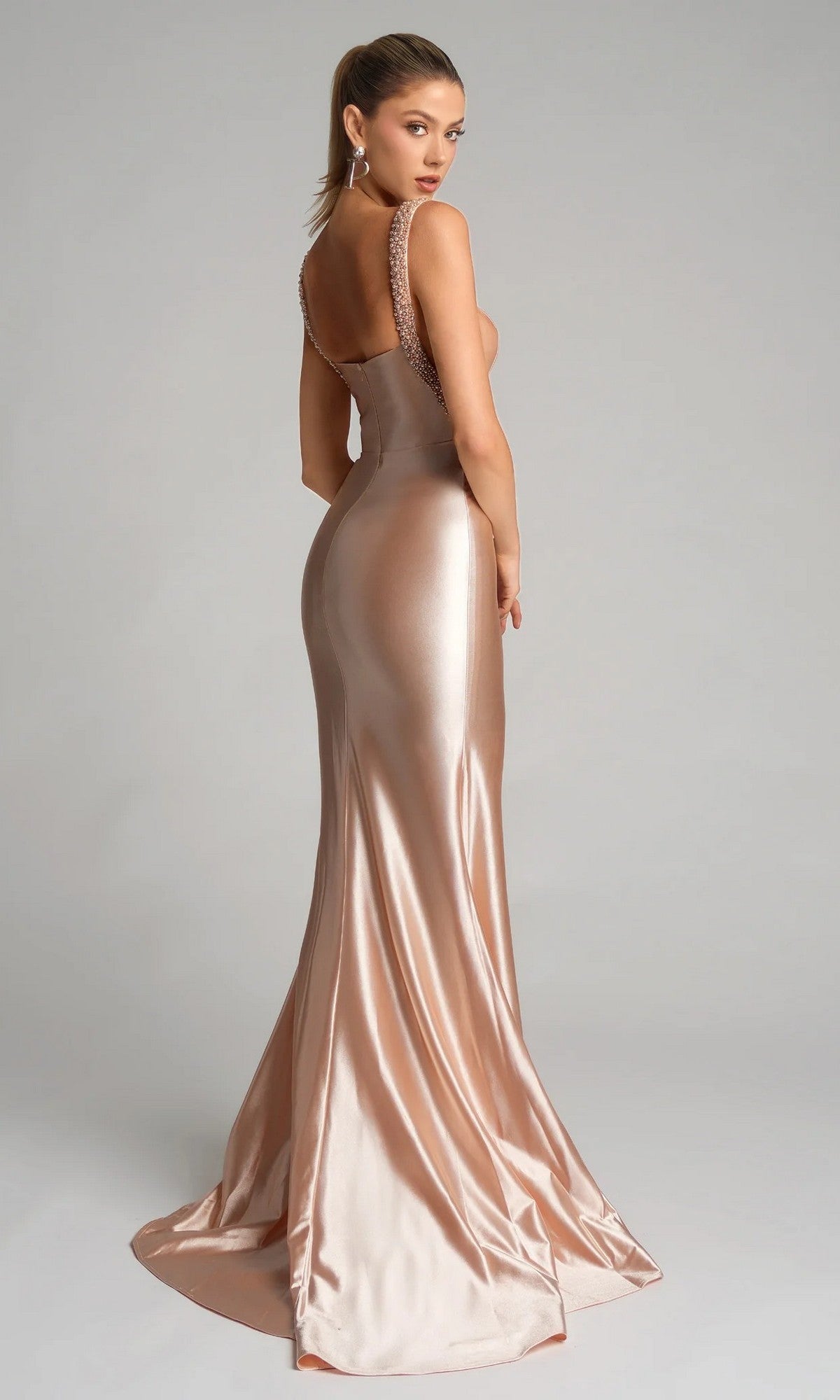 A woman stands facing away in the Portia and Scarlett XO074, a fitted, floor-length metallic rose gold satin formal dress with an open back and beaded shoulder straps, set against a plain light gray background.